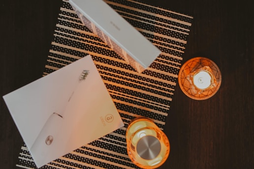 A flat lay composition featuring a boxed electric toothbrush placed on a striped mat. Two lit candles in orange holders create a warm, ambient glow on the dark surface.
