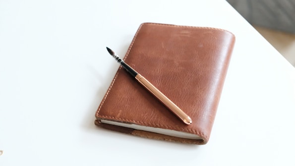 A closed brown leather notebook rests on a light surface, accompanied by a gold-colored fountain pen placed diagonally across its cover.