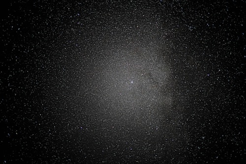 A vast expanse of space sprinkled with countless stars creates a mesmerizing view, with a dense cluster at the center suggesting a galaxy or nebula amid a dark, infinite void.