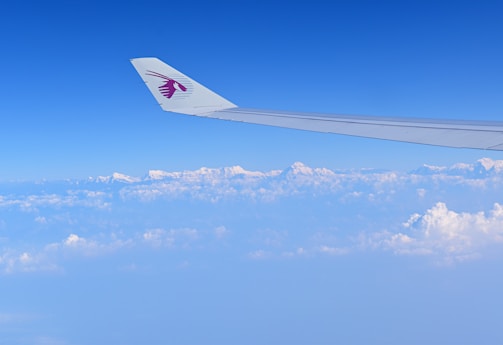 An airplane wing with a logo is visible, soaring high above a layer of fluffy white clouds. In the distance, snow-capped mountain peaks can be seen beneath a vast, clear blue sky.
