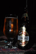 A glass filled with amber-colored beer stands beside a bottle of Tuborg Gold, both placed on a surface with dark ambiance. The bottle is prominently labeled with '%100 Malt' and 'Tuborg Gold'. The soft lighting highlights the golden hue of the beer and reflections on the glass.