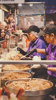 A busy food stall with two people serving noodles from large pans. They wear purple uniforms and black caps, and are assisting customers on the other side of the counter. The atmosphere appears lively, with various food items stacked and customers eagerly waiting.