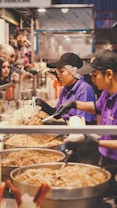 A busy food stall with two people serving noodles from large pans. They wear purple uniforms and black caps, and are assisting customers on the other side of the counter. The atmosphere appears lively, with various food items stacked and customers eagerly waiting.