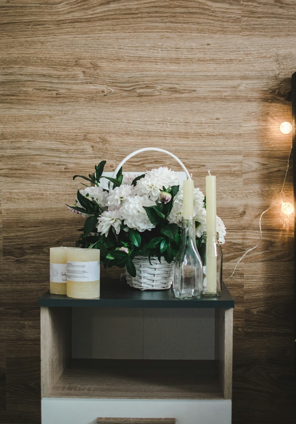 two white candlesticks near white petaled flowers in white wicker basket on white and black wooden end table