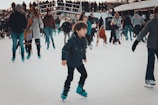 A busy ice skating rink with people of all ages enjoying the winter activity. In the foreground, a young boy is skating confidently while others are either skating or standing around in the background. Many people are dressed warmly in winter clothes, and some are holding hands or supporting each other.