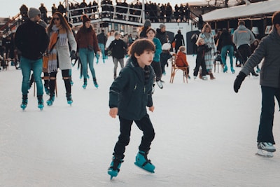A busy ice skating rink with people of all ages enjoying the winter activity. In the foreground, a young boy is skating confidently while others are either skating or standing around in the background. Many people are dressed warmly in winter clothes, and some are holding hands or supporting each other.
