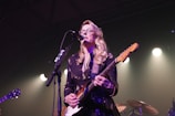 A musician with long blonde hair and glasses is performing on stage, playing an electric guitar and singing into a microphone. Stage lights create a soft, warm glow around the scene.
