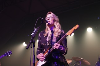 A musician with long blonde hair and glasses is performing on stage, playing an electric guitar and singing into a microphone. Stage lights create a soft, warm glow around the scene.