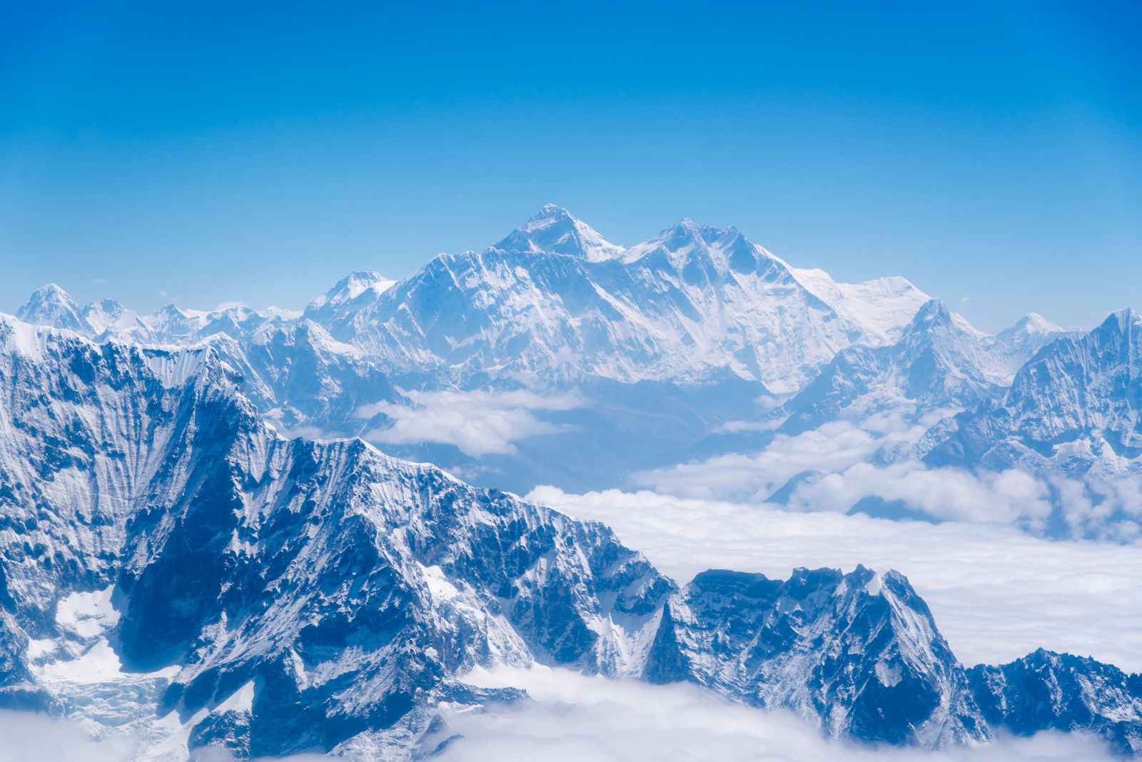 The Everest massif seen from an aircraft, snow ridges rising above a layer of cloud under a clear blue sky