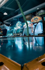 An arcade setting is filled with vibrant lights and game machines. Bright signs and displays feature colorful designs and illuminated lettering, creating an energetic atmosphere. The surface reflects the neon colors, adding to the dynamic and playful environment.