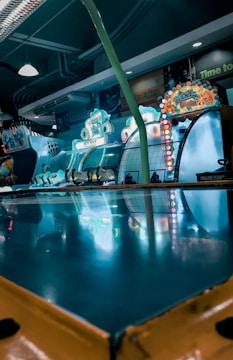 An arcade setting is filled with vibrant lights and game machines. Bright signs and displays feature colorful designs and illuminated lettering, creating an energetic atmosphere. The surface reflects the neon colors, adding to the dynamic and playful environment.