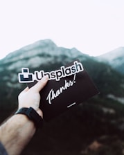 A hand holds a card with the words 'Unsplash Thanks!' against a blurred background of mountains and sky.