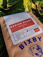 A package of coffee labeled 'Medium Roast' with details including its origin, Kenya and Guatemala, and a roast date. The packaging features a graphic of a dog and the brand name 'Bixby'. The background contains vibrant green leaves.