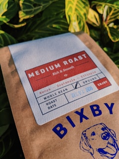 A package of coffee labeled 'Medium Roast' with details including its origin, Kenya and Guatemala, and a roast date. The packaging features a graphic of a dog and the brand name 'Bixby'. The background contains vibrant green leaves.