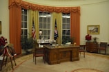 A spacious, elegantly decorated office with an ornate wooden desk at the center, featuring various items and framed pictures on it. Large windows with gold and red curtains flank two flags standing upright. The room has potted plants and a wooden chest of drawers with a painting above it. Red floral arrangements and a saddle with decorative elements are also visible.