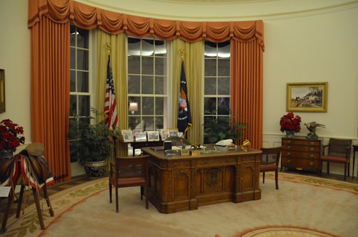 A spacious, elegantly decorated office with an ornate wooden desk at the center, featuring various items and framed pictures on it. Large windows with gold and red curtains flank two flags standing upright. The room has potted plants and a wooden chest of drawers with a painting above it. Red floral arrangements and a saddle with decorative elements are also visible.