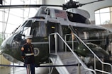 A military helicopter is displayed indoors, featuring an American flag and a presidential seal. A mannequin dressed in a United States Marine Corps uniform stands near the entrance of the helicopter, which has a small staircase with a welcome sign.