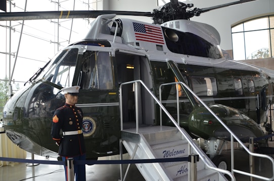 A military helicopter is displayed indoors, featuring an American flag and a presidential seal. A mannequin dressed in a United States Marine Corps uniform stands near the entrance of the helicopter, which has a small staircase with a welcome sign.