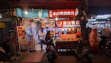 An evening scene at a bustling street market featuring a food stall with illuminated signage and a variety of goods displayed. People are gathered around, including a person in a white jacket engaging with a nearby claw machine. Scooters are parked in front of the stalls, adding to the vibrant, lively atmosphere.
