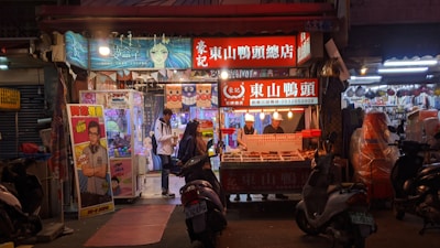 An evening scene at a bustling street market featuring a food stall with illuminated signage and a variety of goods displayed. People are gathered around, including a person in a white jacket engaging with a nearby claw machine. Scooters are parked in front of the stalls, adding to the vibrant, lively atmosphere.