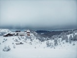 A snow-covered landscape with a large resort or lodge nestled among snow-laden trees and hills. The sky is overcast with thick clouds, creating a serene yet cold atmosphere. Buildings are set against a backdrop of distant hills, partially obscured by the foggy haze.