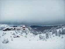 A snow-covered landscape with a large resort or lodge nestled among snow-laden trees and hills. The sky is overcast with thick clouds, creating a serene yet cold atmosphere. Buildings are set against a backdrop of distant hills, partially obscured by the foggy haze.