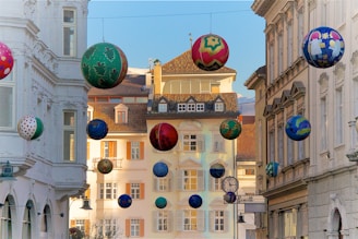 A street scene with decorative, colorful balls suspended in the air between buildings. The buildings feature classic European architectural details with white facades and large windows. The decorations vary in design and color, adding a festive and whimsical touch to the scene.