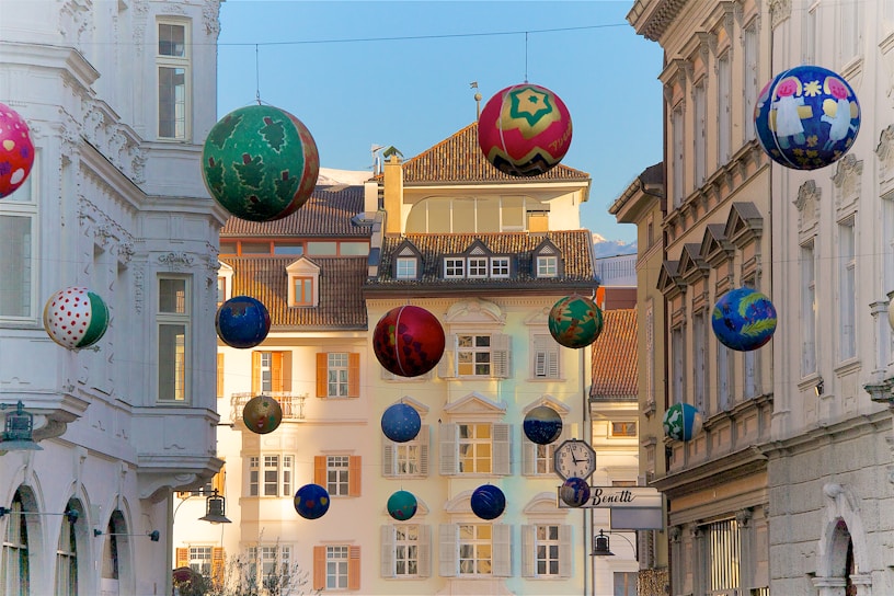 A street scene with decorative, colorful balls suspended in the air between buildings. The buildings feature classic European architectural details with white facades and large windows. The decorations vary in design and color, adding a festive and whimsical touch to the scene.