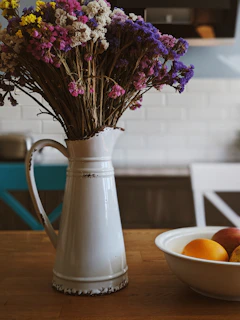 A vibrant ceramic pitcher in sunset orange sitting beside a vase of wildflowers on a rustic kitchen shelf