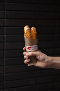 A hand holding a paper sleeve containing two crispy, orange-brown cheesesticks with melted cheese visible. The sleeve is branded with a label that reads 'Mister Burger'. The background consists of a dark brick wall.