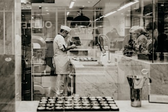 A chef wearing a white uniform and hat is standing in a professional kitchen, likely baking. The environment is clean and organized, with marble counters and kitchen equipment visible. Several trays of pastries are on the counter, and the chef seems focused on their task. Other people are nearby, possibly assisting or observing.