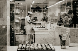 A chef wearing a white uniform and hat is standing in a professional kitchen, likely baking. The environment is clean and organized, with marble counters and kitchen equipment visible. Several trays of pastries are on the counter, and the chef seems focused on their task. Other people are nearby, possibly assisting or observing.