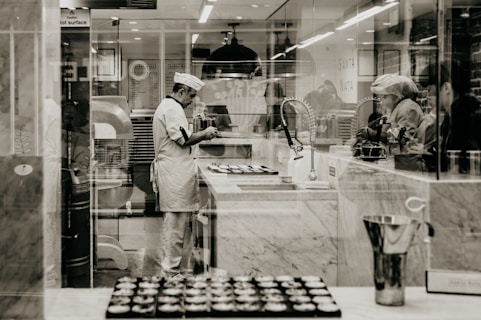 A chef wearing a white uniform and hat is standing in a professional kitchen, likely baking. The environment is clean and organized, with marble counters and kitchen equipment visible. Several trays of pastries are on the counter, and the chef seems focused on their task. Other people are nearby, possibly assisting or observing.