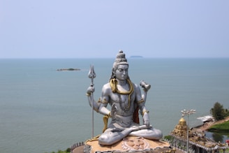 A large statue of a deity is seated in a meditative pose near the sea with a clear, blue sky overhead. The statue holds a trident in one hand and is adorned with multiple necklaces. Nearby, a golden temple-like structure is visible along the coastline.