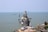 A large statue of a deity is seated in a meditative pose near the sea with a clear, blue sky overhead. The statue holds a trident in one hand and is adorned with multiple necklaces. Nearby, a golden temple-like structure is visible along the coastline.