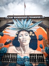 A large mural painted on the side of a building that reads 'Tucson Warehouse and Transfer Co.' The mural depicts a woman with long dark hair adorned with yellow flowers and a large agave plant on her head. The background features an orange and blue color scheme with abstract shapes and cactus elements. An American flag is visible on top of the building.