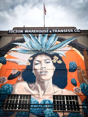 A large mural painted on the side of a building that reads 'Tucson Warehouse and Transfer Co.' The mural depicts a woman with long dark hair adorned with yellow flowers and a large agave plant on her head. The background features an orange and blue color scheme with abstract shapes and cactus elements. An American flag is visible on top of the building.