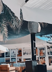 A stylish outdoor seating area with woven wicker lampshades hanging from a canopy. Palm fronds partially shade the space, adding to the tropical ambiance. Photographs are displayed on the walls of the surrounding building, featuring various scenes and people.