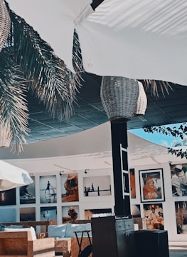 A stylish outdoor seating area with woven wicker lampshades hanging from a canopy. Palm fronds partially shade the space, adding to the tropical ambiance. Photographs are displayed on the walls of the surrounding building, featuring various scenes and people.