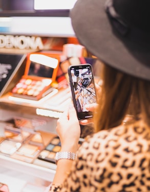 A person wearing a hat and a patterned shirt is holding a smartphone, capturing a photo of a makeup display in a store. The display features various palettes and cosmetic products arranged neatly in bright and inviting packaging.