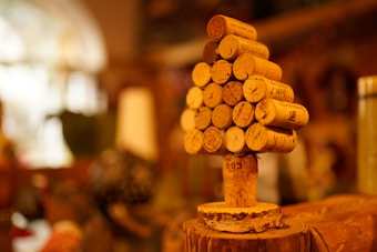 A small tree-like structure is crafted from wine corks, stacked in a conical shape on top of a single vertical cork. The background is softly blurred, featuring warm colors that suggest an indoor setting, possibly in a cozy room or a wine bar.