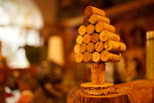 A small tree-like structure is crafted from wine corks, stacked in a conical shape on top of a single vertical cork. The background is softly blurred, featuring warm colors that suggest an indoor setting, possibly in a cozy room or a wine bar.