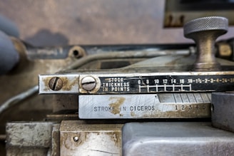 An industrial metal machine component displaying a scale for measuring thickness in points, alongside measurements labeled 'STROKE IN CICEROS'. The device has a metallic knob and visible screws, with some wear and signs of aging on the metallic surface.
