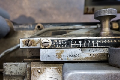 An industrial metal machine component displaying a scale for measuring thickness in points, alongside measurements labeled 'STROKE IN CICEROS'. The device has a metallic knob and visible screws, with some wear and signs of aging on the metallic surface.