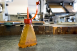 An oil can with a red nozzle and cap sits on a metal surface in a workshop or industrial setting. The bottle is transparent with an amber hue and has a number '24' marked on it. The background features blurred machinery and equipment, suggesting an environment related to manufacturing or maintenance.