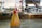 An oil can with a red nozzle and cap sits on a metal surface in a workshop or industrial setting. The bottle is transparent with an amber hue and has a number '24' marked on it. The background features blurred machinery and equipment, suggesting an environment related to manufacturing or maintenance.