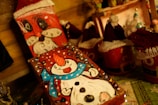 A festive scene featuring handmade crafts, including painted wooden blocks with snowman and Santa Claus designs. The snowman is adorned with a red hat and a blue scarf, while Santa is depicted with his iconic white beard. Surrounding these are small red containers with knitted hats, resembling Santa hats. The warm lighting adds a cozy atmosphere to the display.