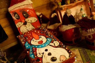 A festive scene featuring handmade crafts, including painted wooden blocks with snowman and Santa Claus designs. The snowman is adorned with a red hat and a blue scarf, while Santa is depicted with his iconic white beard. Surrounding these are small red containers with knitted hats, resembling Santa hats. The warm lighting adds a cozy atmosphere to the display.