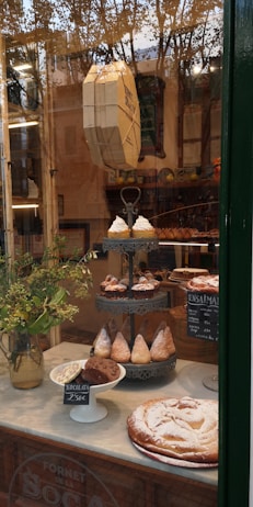 A bakery window display featuring an assortment of pastries, including a variety of croissants and sweet treats stacked on a decorative stand. The setting includes a vase with green foliage and visible reflections of trees and buildings outside. There are handwritten chalkboard signs in Spanish with prices.