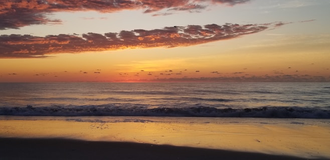 A serene ocean scene at sunrise with calming waves gently hitting the shore. The sky is painted in warm hues of orange, yellow, and pink with scattered clouds creating a picturesque and tranquil ambiance.